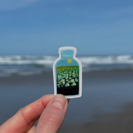 A hand holding a clear vinyl sticker with a daisy terrarium design, with a beach and ocean in the background.