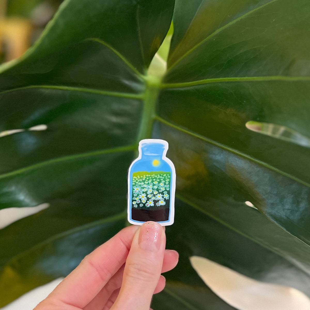 Hand holding a small bottle with plants sticker against a leafy green background
