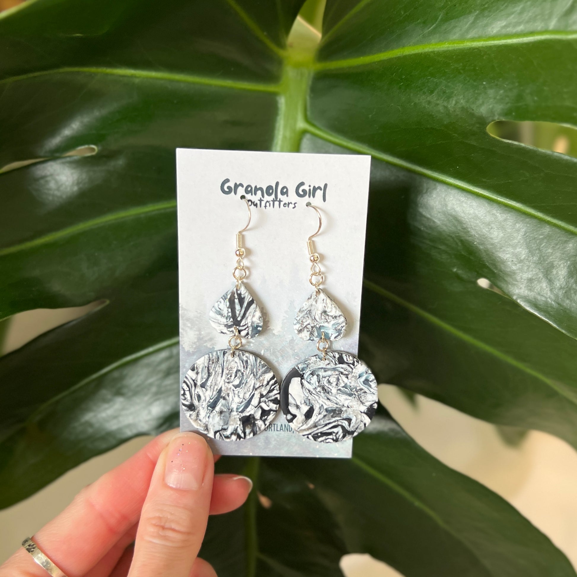 Abstract earrings on a 'Grande Girl' branded card held against a green leafy background