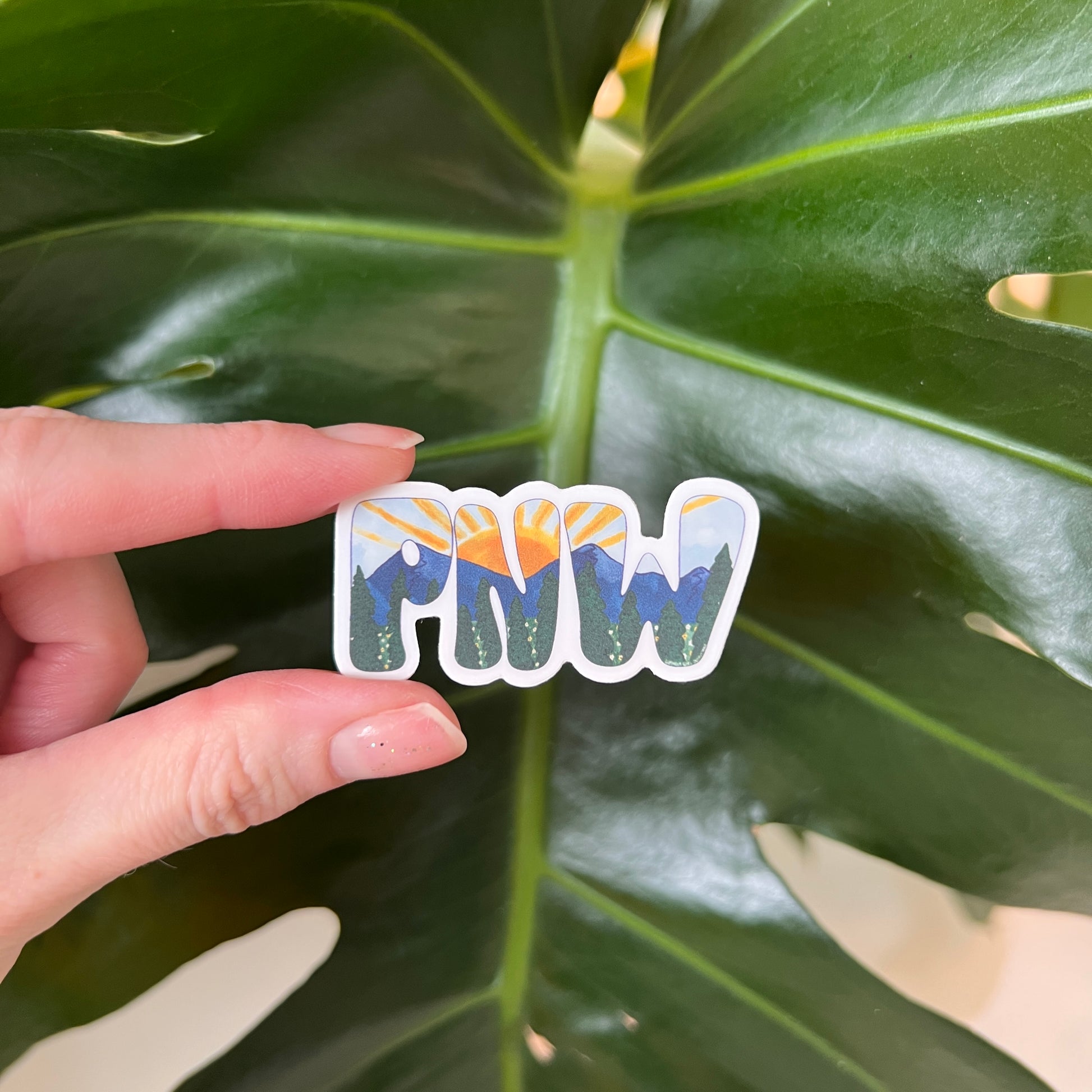 fingers holding a PNW sticker against a leafy background