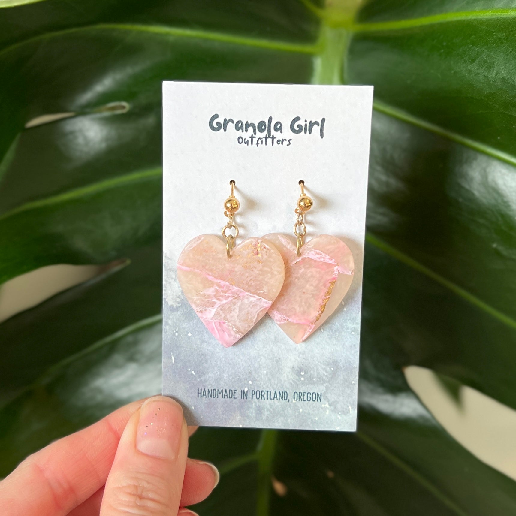 Heart-shaped earrings on a 'Granola Girl Outfitters' card held against a leafy green background.
