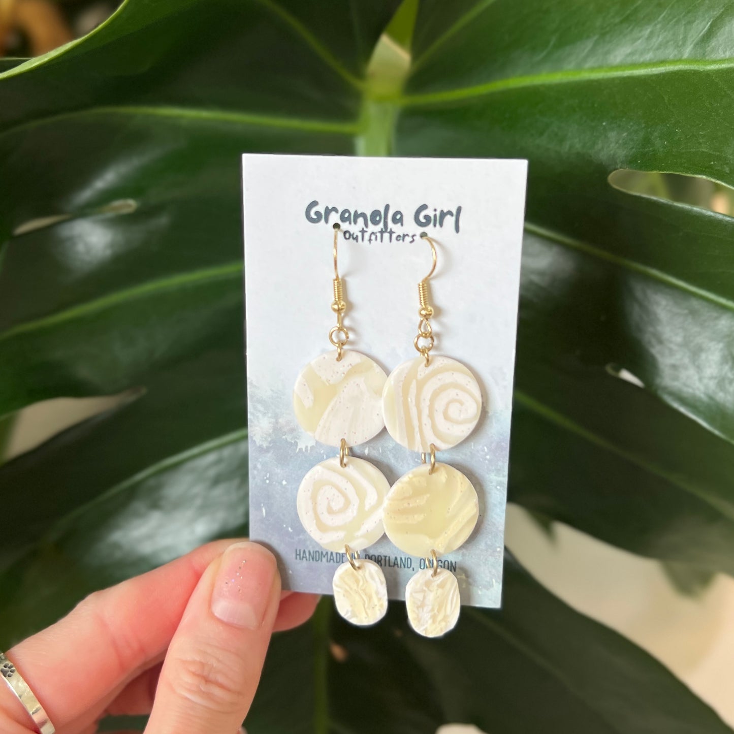 Pair of earrings on a 'Granola Girl Outfitters' card held in front of green leaves.