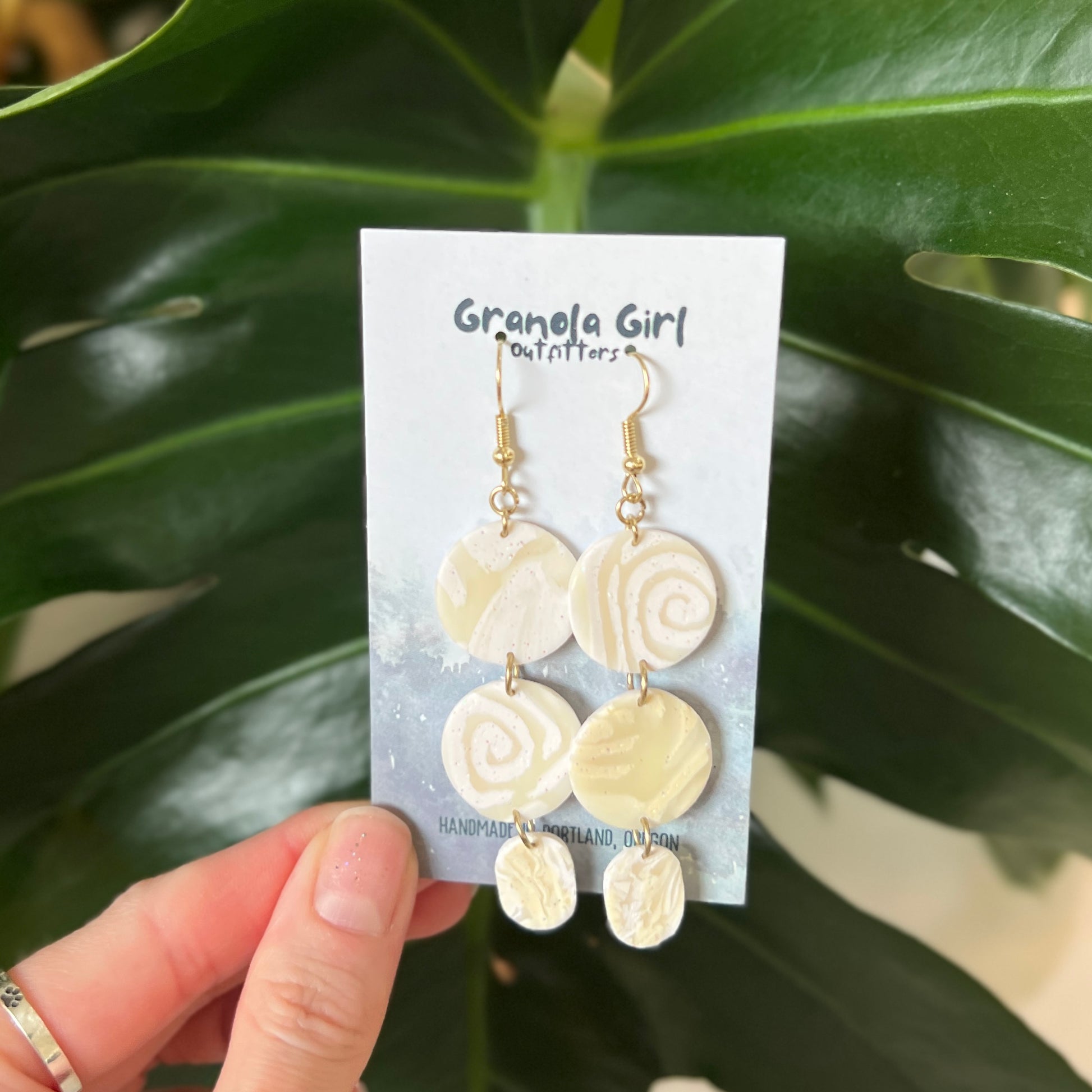Pair of earrings on a 'Granola Girl Outfitters' card held in front of green leaves.