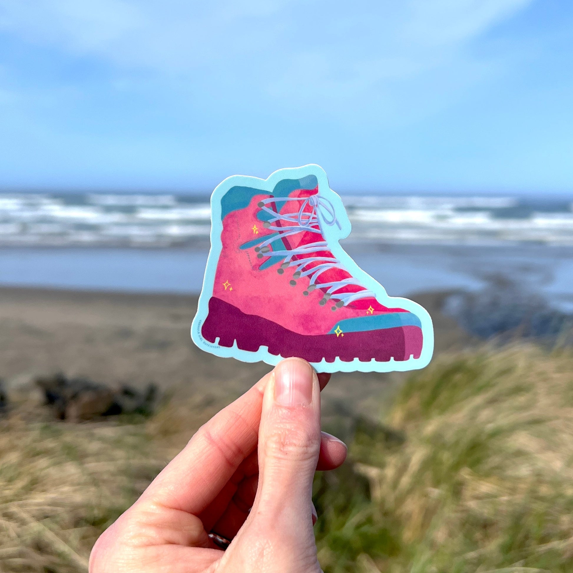 A hand holding a sticker in the shape of a hiking boot with a colorful design, with a beach and grass in the background.