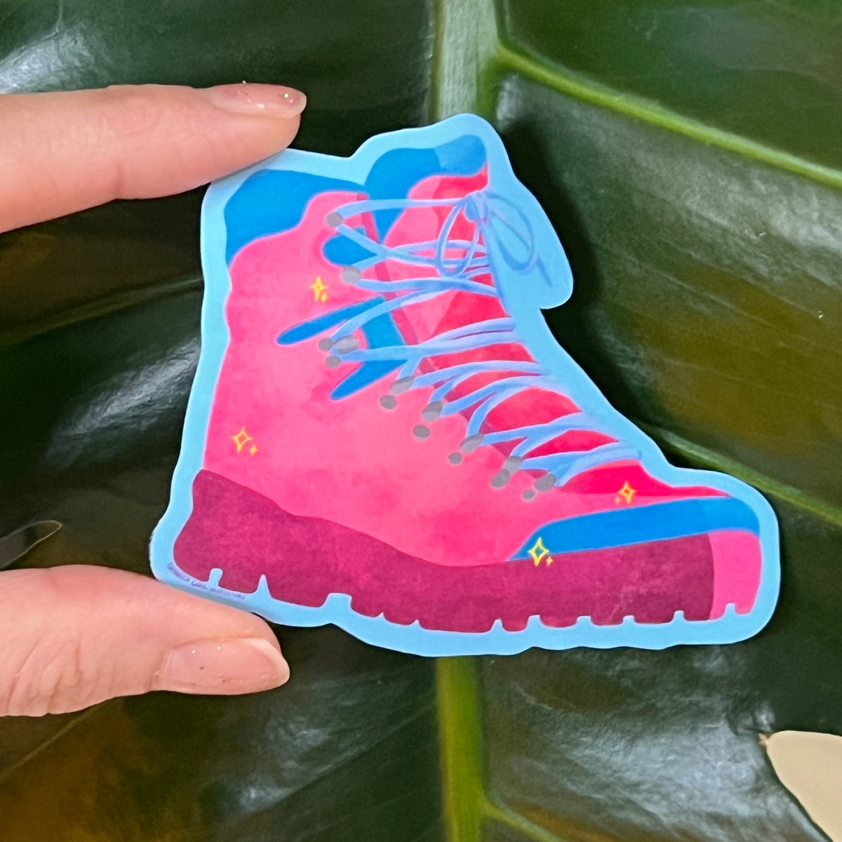 Hand holding a pink and blue boot-shaped sticker against a green leafy background
