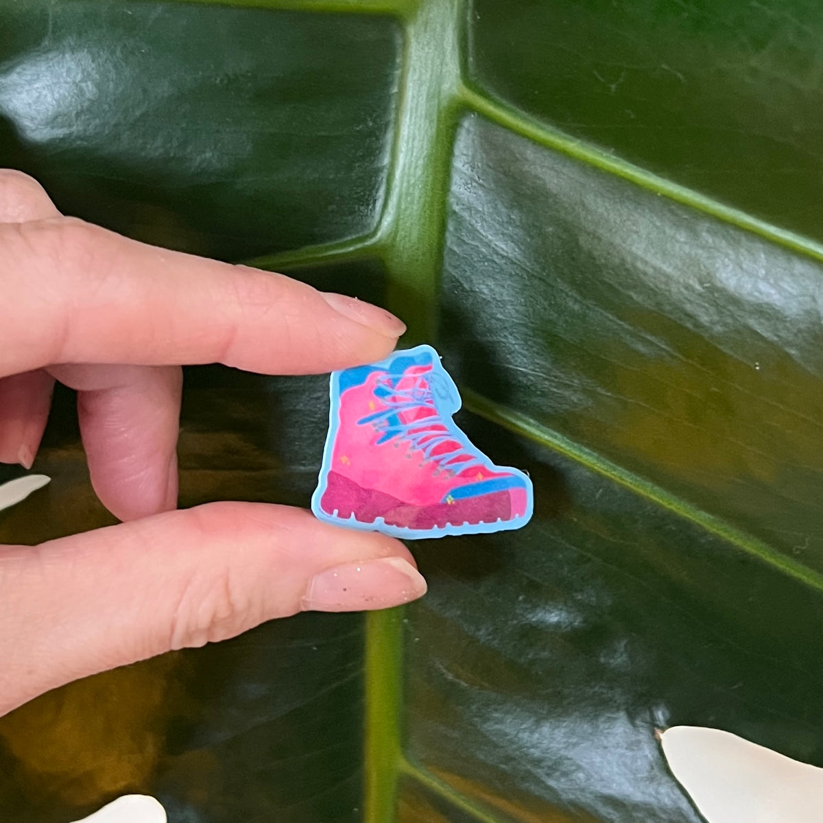 Hand holding a colorful boot-shaped sticker against a green leaf background