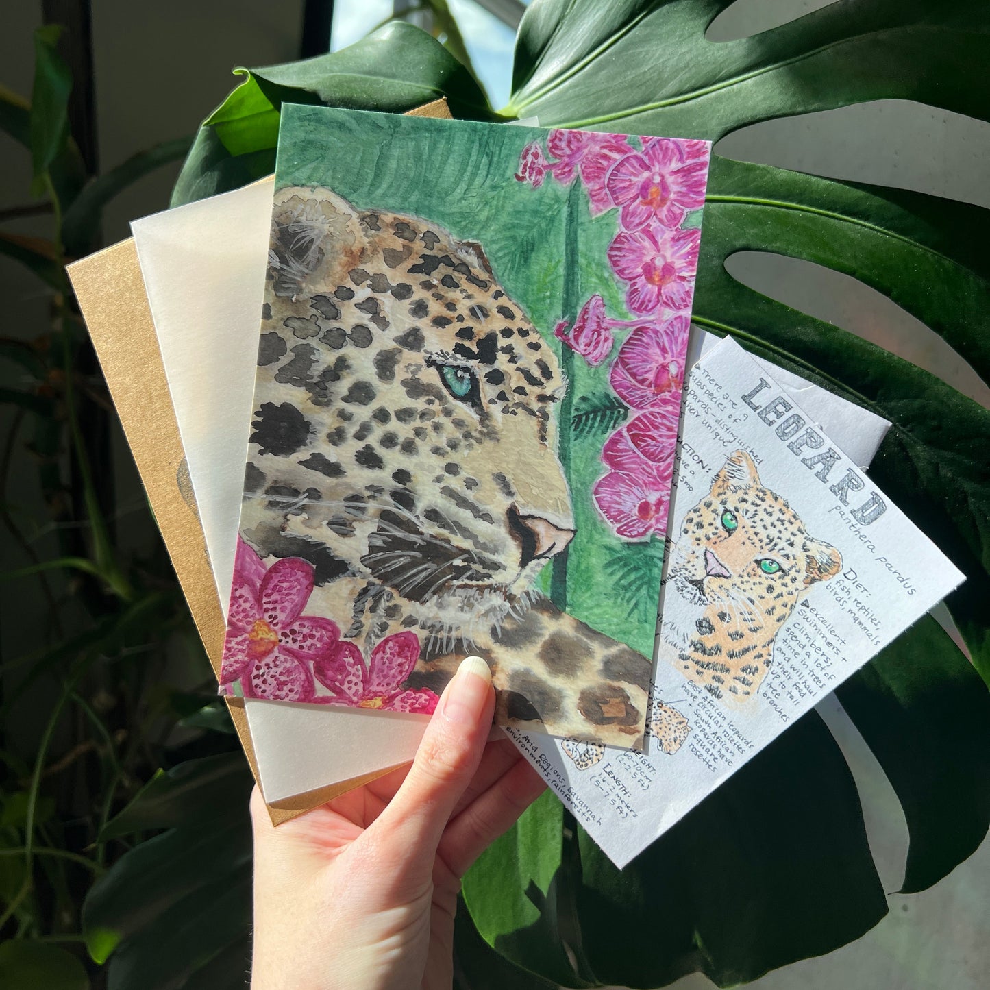 art print of a leopard and other paper items being held up by a hand and green plants in the background