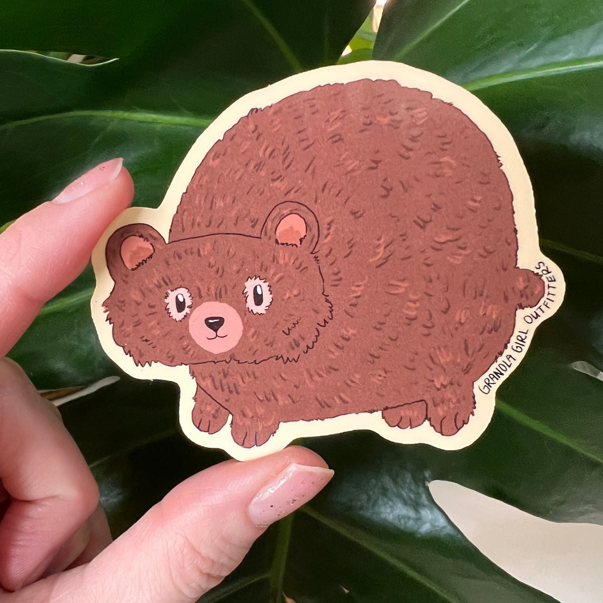 Cartoon bear sticker held by a hand against a leafy green background