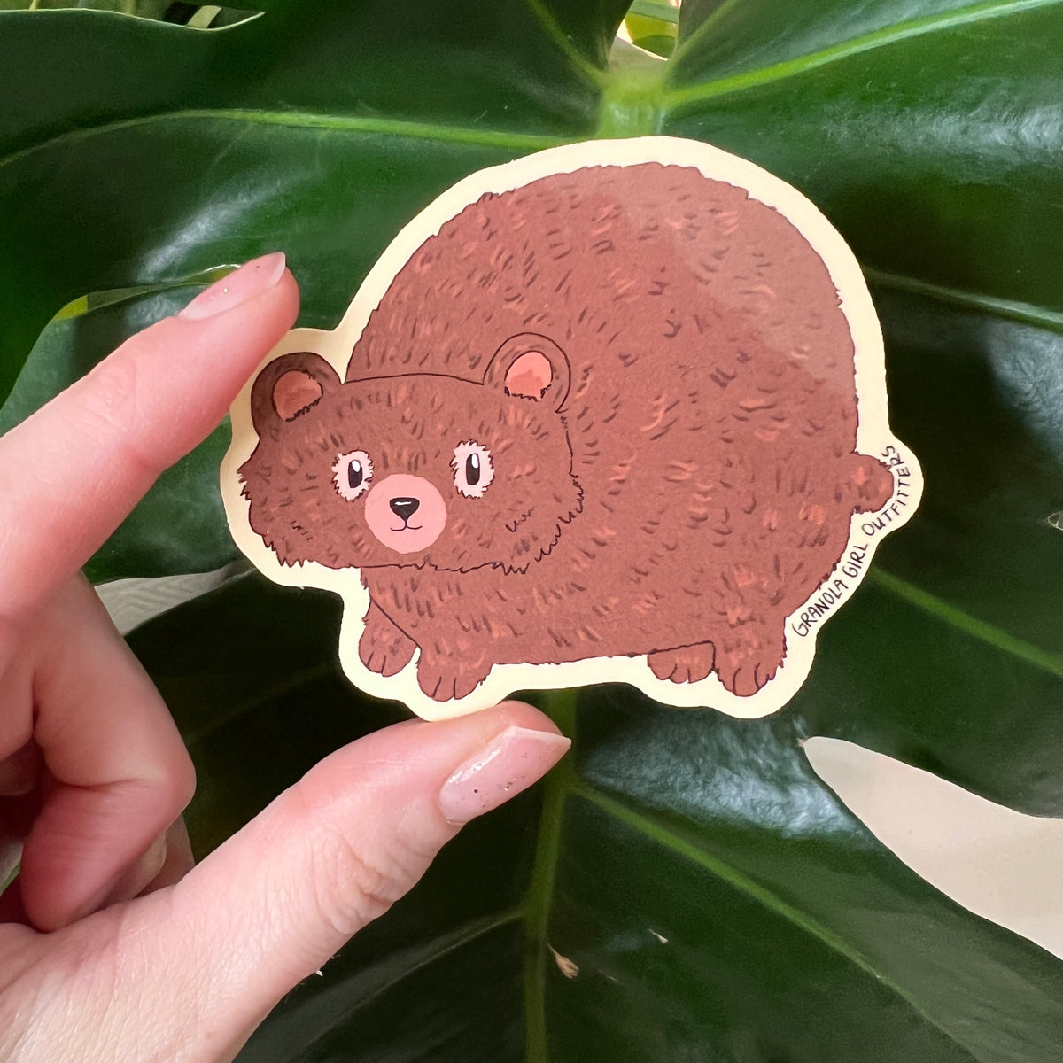 Hand holding a cartoon bear sticker against a leafy green background