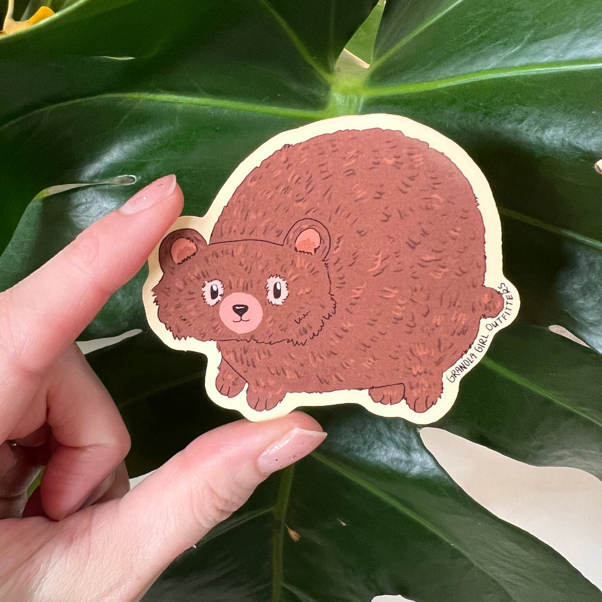 Hand holding a bear-shaped sticker against a leafy green background