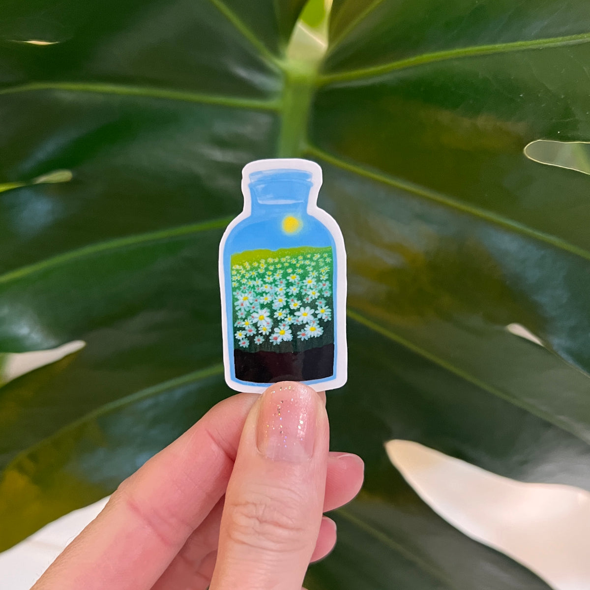 Small bottle-shaped sticker with a floral design held between fingers against a leafy green background