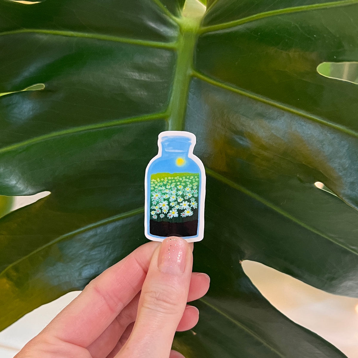 Hand holding a sticker of a bottle with flowers against a leafy background