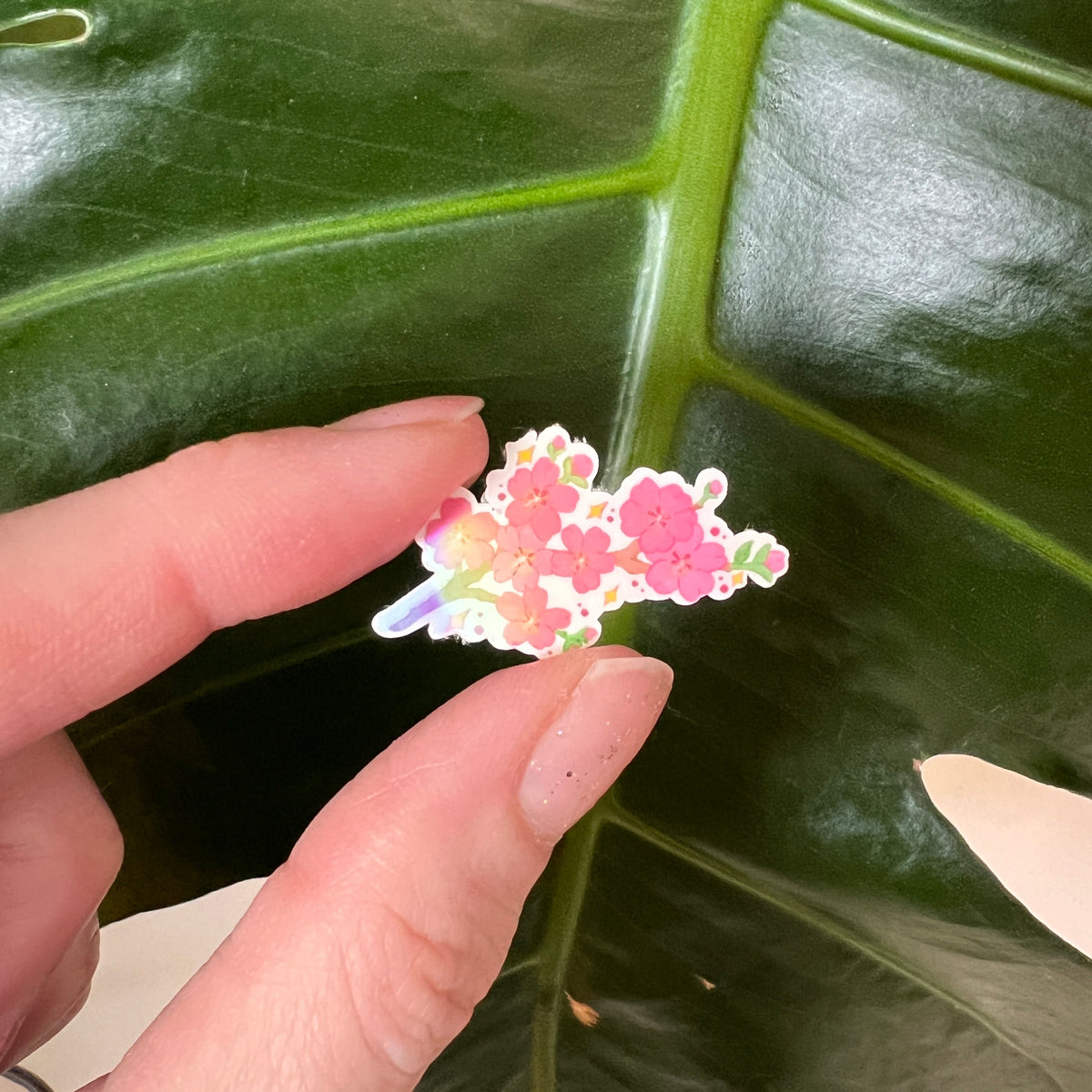 Hand holding a colorful flower sticker against a green leaf background