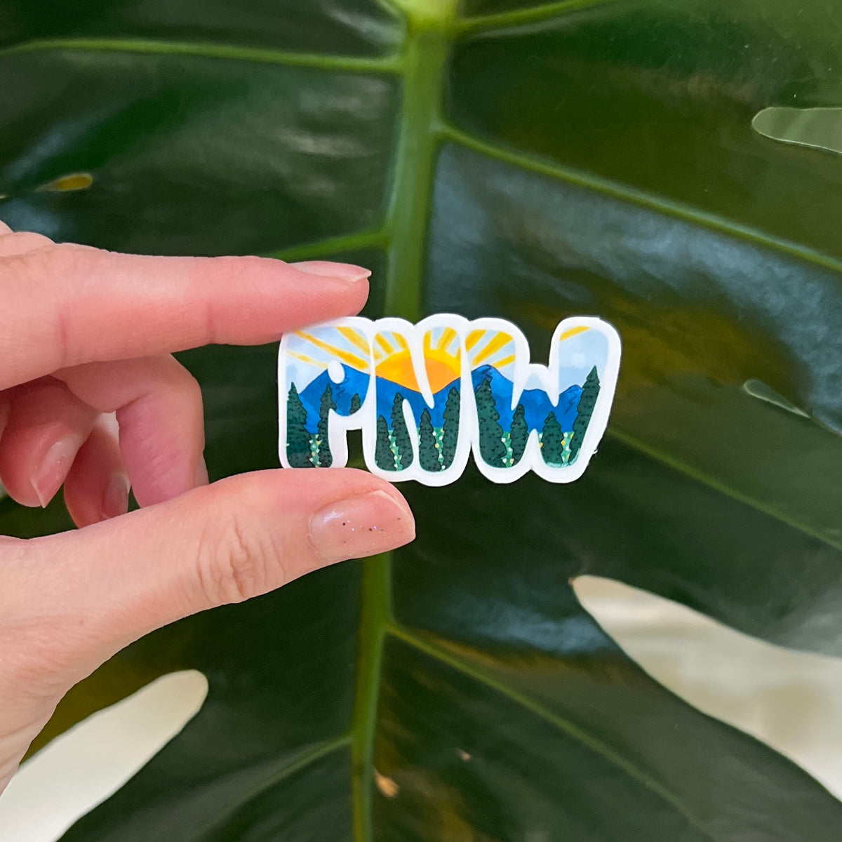 Hand holding a nature-themed sticker with mountains and sun on a leaf background
