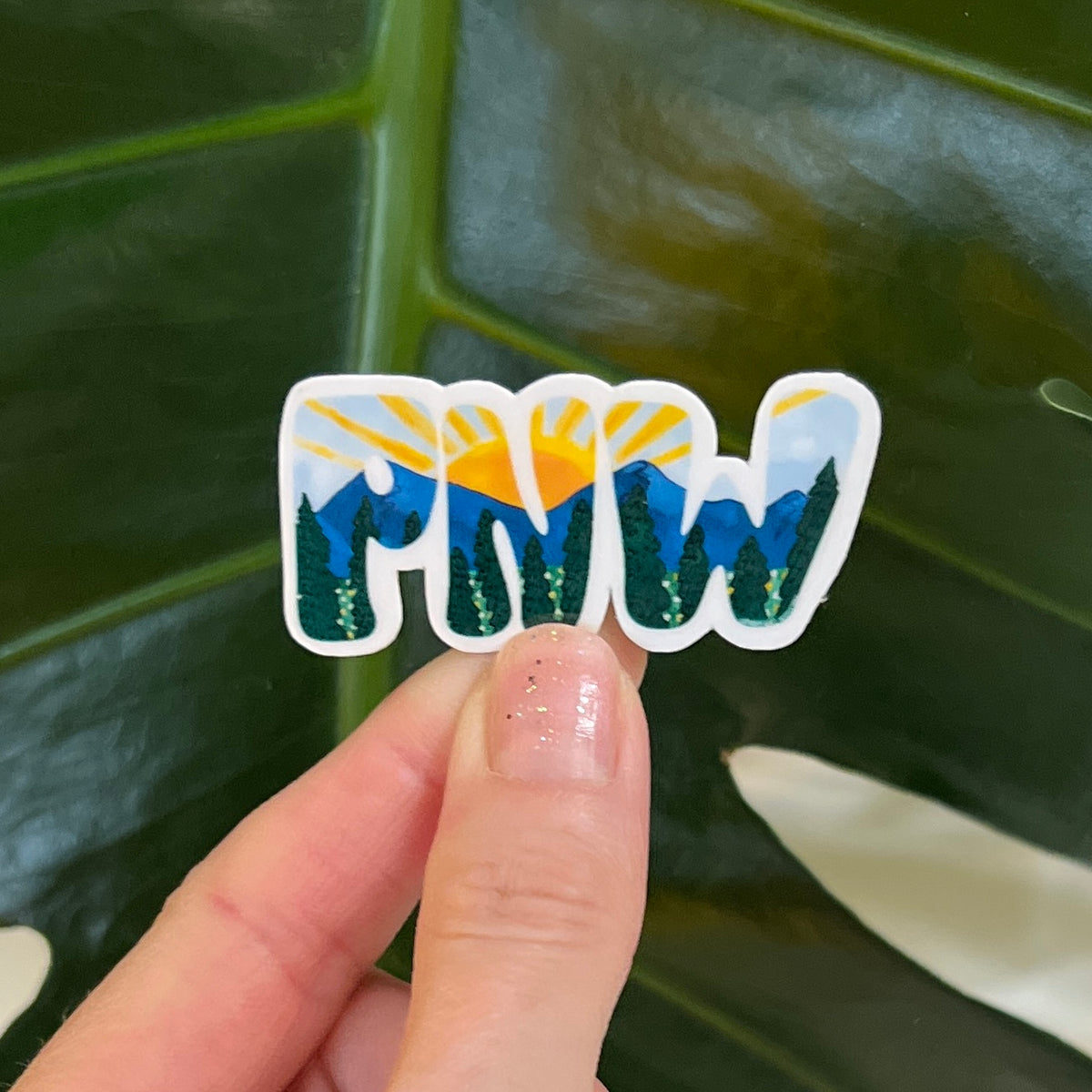 Hand holding a sticker with 'PNW' design against a leafy background