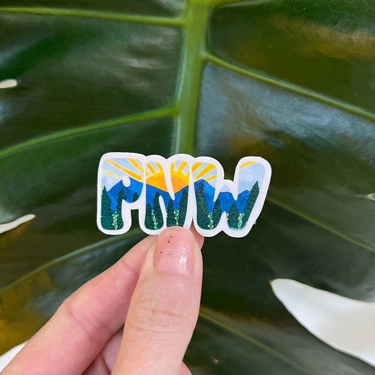 Hand holding a sticker with 'PNW' design against a leafy background