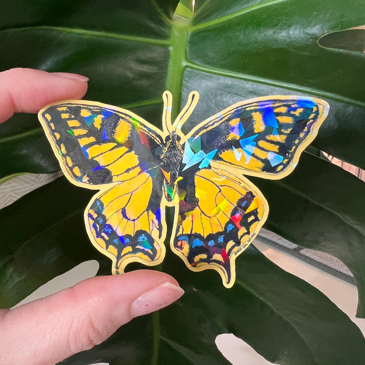 Colorful butterfly-shaped sticker held by a hand against a leafy background